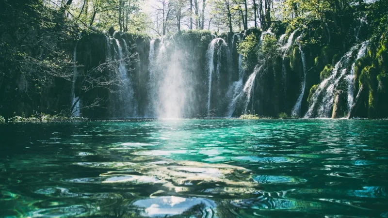 A beautiful multi-tiered waterfall cascading into a turquoise lake surrounded by lush green forest.